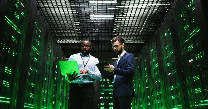 Mixed-races men with laptop computer and tablet device. Software specialists walking at servers and checking big data information. Multiethnic males analytics colleagues discussing digital security.