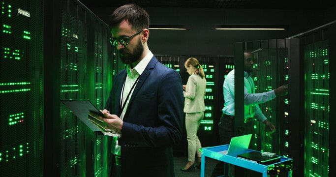 Caucasian man technician in database room with servers, tapping on tablet device and coding information. Male security worker in datacenter using gadget. African American cryptograph on background.