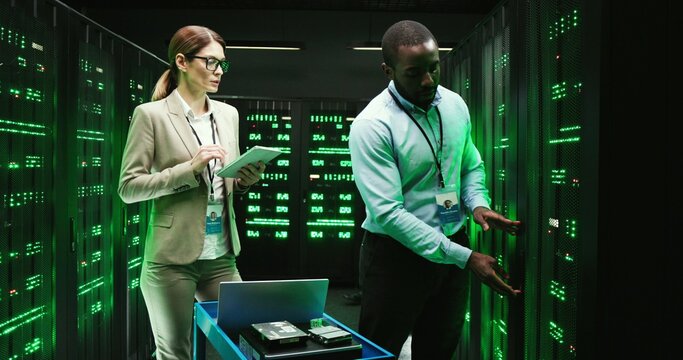 African American man checking server and Caucasian woman tapping on tablet device. Software engineers programming big data processors. Male and female analytics working on datacenter security.