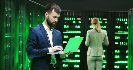 Caucasian young man, software specialist at servers with laptop computer, typing on keyboard and checking big data processors. Female analytic working on digital information security on background.