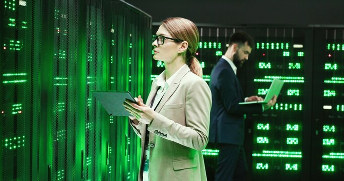Caucasian woman standing at server with tablet device, tapping and checking worker server. Female analytic in data storage working on digital security Male colleague with laptop computer on background