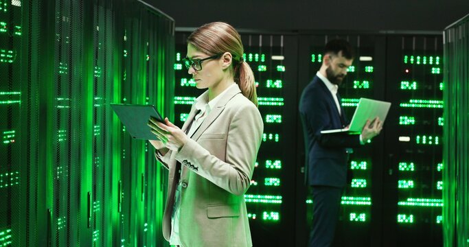 Caucasian woman standing at server with tablet device, tapping and checking worker server. Female analytic in data storage working on digital security Male colleague with laptop computer on background - Powered by Adobe