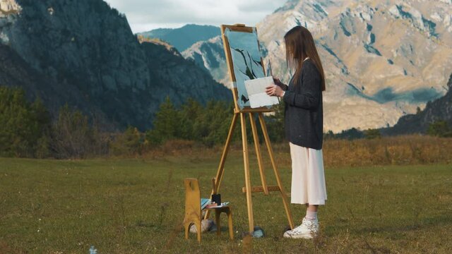 Beautiful girl paints a picture in the mountains. A professional artist creates a landscape on a molbert. Hobbies, hobbies and work in the fresh air.