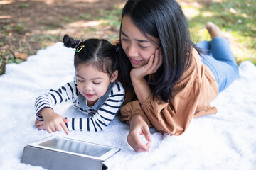 Closeup daughter with her mom using tablet and lying on the ground in the public garden. Girl playing game from the tablet. A good relationship at the weekend. Education, online and family concept