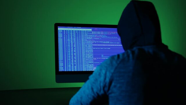 Back view of hacker programmer wearing a hood, writing green code in a computer screen. Unrecognizable man hacking in a dark room. Concept dark internet, cyber security.