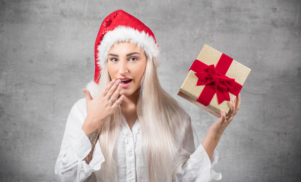 Surprised beautiful blond woman with wears christmas santa claus hat - cap and holding red gift box - present with ribbon, isolated on gray background