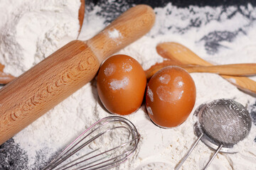 A mound of white flour on a dark surface. Nearby are kitchen appliances for cooking and two chicken eggs.