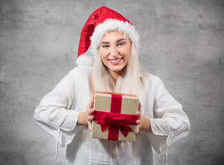 Santa woman shows christmas gift isolated on gray background. Happy young girl wearing red santa hat and holding present box