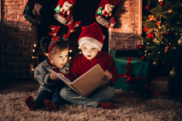 Brother and sister are sitting and reading a book. Christmas mood. Children in New Year's costumes against the background of a Christmas tree and a fireplace