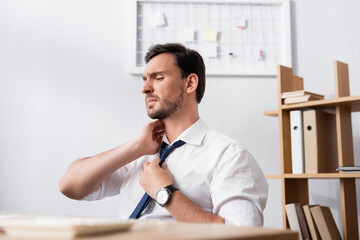 Businessman with painful neck sitting at workplace with blurred desk on foreground
