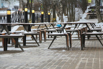 Tables and benches covered with snow stand on the city street on a pavement next to the restaurant...