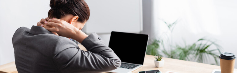 Back view of female office worker with hurting neck sitting at desk with laptop on blurred background, banner