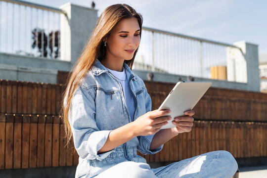 Attractive woman in denim clothes sits on park bench, holds smart tablet in her hands. Portrait of beautiful girl