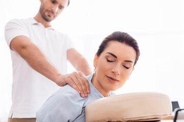 Focused masseur doing shoulders massage of positive businesswoman with closed eye on blurred background