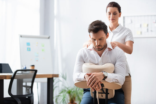 Smiling masseuse massaging shoulders of businessman sitting on massage chair in office on blurred background