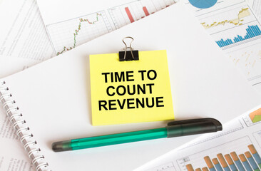 A yellow sticker with text Time To Count Revenue is in a Notepad with a green pen financial charts and documents