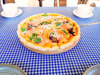 Fresh pizza with red fish greens and cheese stands on the table with a checkered tablecloth and two white cups