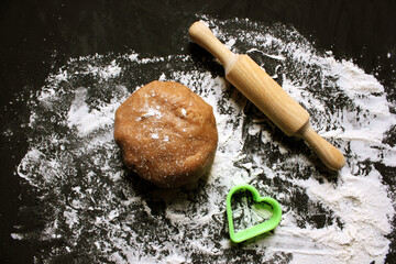 Dough, rolling pin and shape for cookies in the form of a heart on a black background. Ginger cookie dough on a floured black surface. View from above. The concept of cooking. Copying space for text