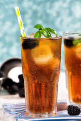 The glass of iced tea with blackberry on wooden table. Cuba Libre or long island cocktail, cold drink or lemonade with lemon and basil. Green turquoise on background