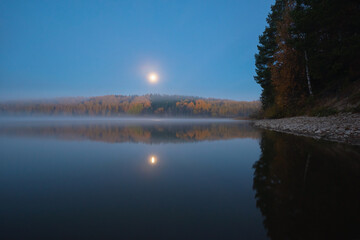 Obraz premium Scenic view of the river banks with trees covered in autumn colored leafs and mist during sunrise and full moon set
