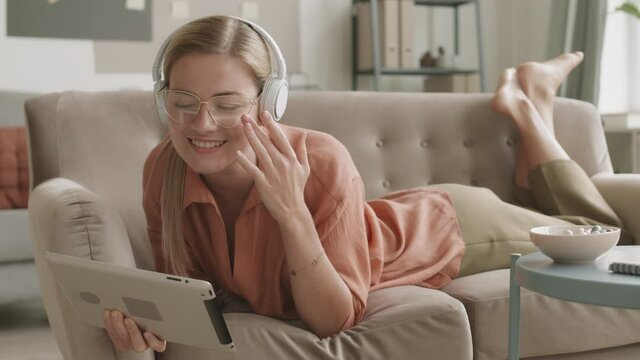 Full shot of young Caucasian woman lying down on stomach on couch barefoot, having video call using tablet and wireless over-head earphones. Girl waving hand, speaking, smiling eating dragee