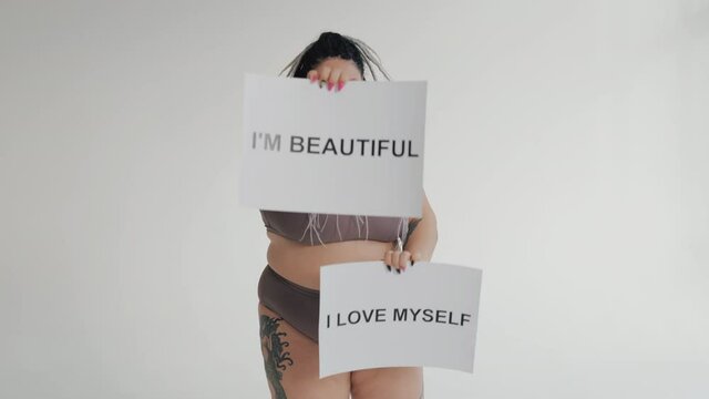 Happy fat woman in underwear with drearlocks dancing with body positive posters. Diverse beauty, diversity tolerance concept. Stout girl in sweamwer isolated smiling to camera.