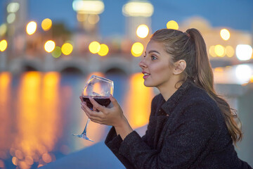 A girl with a beautiful smile is on the embankment and holding a glass of red wine. evening city in the background.