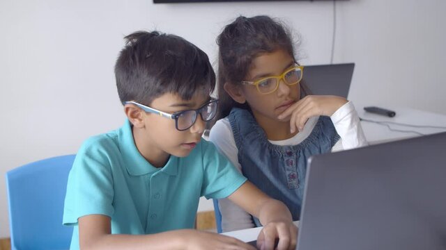 Couple of schoolchildren in glasses working on task together, using laptop in classroom, watching content and laughing. Static shot. Digital education concept
