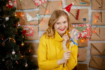 happy smiling girl in red christmas hat with lollipop near decorated christmas tree