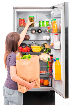 A girl in casual clothes takes food out of a paper bag and puts it in a refrigerator with dark-colored doors. Vertical photo, white background, rear view.