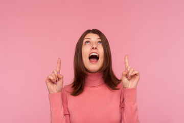 Extremely surprised happy woman with brown hair in pink sweater pointing fingers up on empty space, satisfied with advertisement area. Indoor studio shot isolated on pink background