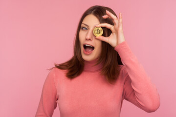 Happy cheerful brunette woman in pink sweater having fun holding crypto coin near her eye and winking looking at camera, bitcoin cryptocurrency. Indoor studio shot isolated on pink background