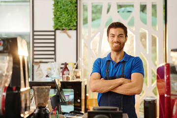 Portrait of handsome smiling coffeeshop barista folding arms and looking at camera