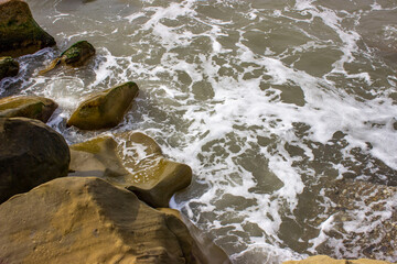Stormy sea. Powerful big foam waves beat against big beautiful rocks, rocks, breakwaters. A scattering of spray. Cloudy weather with bright light.