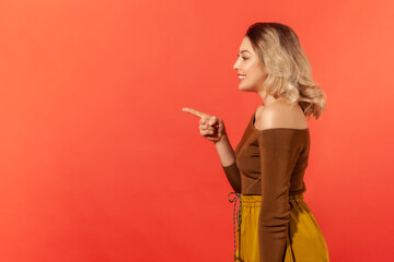 Side view portrait of beautiful toothy smiling woman with blonde hair in brown blouse pointing finger to the left, looking with astonishment. Indoor studio shot isolated on red background