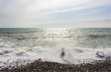 Stormy sea. Gray-blue, cloudy. Foam waves beat on the shore, rocks, breakwaters. Cloudy weather. Sunlight reflects in the water and flickers.