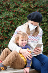 Mother with child on park bench, using mobile phone, Woman with protective mask, coronavirus prevention