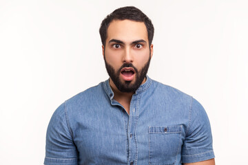 Fototapeta premium Surprised wondered man with beard in blue shirt looking at camera with big eyes and open mouth, shocked with unexpected news. Indoor studio shot isolated on white background
