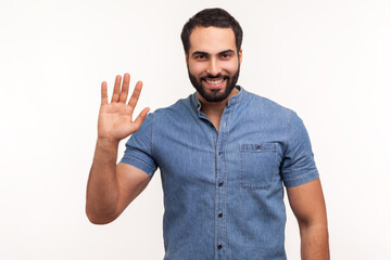 Fototapeta premium Friendly positive bearded man in blue shirt greeting you rising hand and waving, saying hi, glad to see you, looking at camera with toothy smile. Indoor studio shot isolated on white background