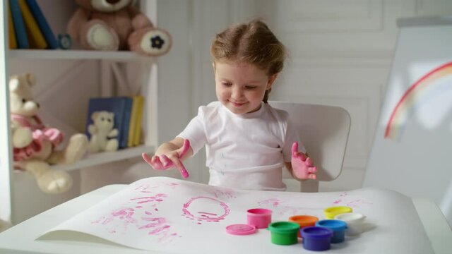 Lovely happy little girl preschooler uses fingers covered in pink paint to create cute abstract images on big sheet of paper. Kid spending time at home with pleasure.