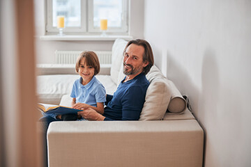 Adorable kid and his bearded dad resting on sofa