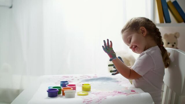 Cute three years old kid enjoys finger painting activity, sits at table with dirty hands covered in multicolored paint. Little girl having fun at home.