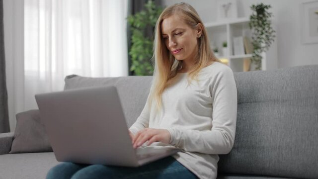 Beautiful blonde relaxing on grey sofa and using modern laptop. Happy lady in domestic outfit holding wireless computer in knees during free time at home.