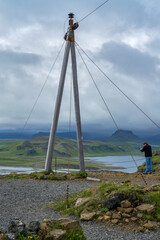 A slender tripod structure with wires stands on a grassy hill in Dyrhólaey, Iceland, against a...