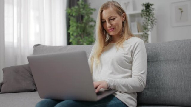 Smiling woman with blond hair sitting on cozy sofa and typing on portable laptop. Happy female using wireless gadget while staying at home.