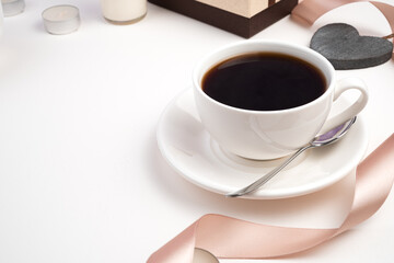 Coffee Cup on saucer with spoon on holiday background with gift box, ribbon and heart on light background. Side view with space for copying.