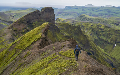 A lone hiker follows a narrow ridge trail in Iceland's Thakgil area, surrounded by dramatic mossy...