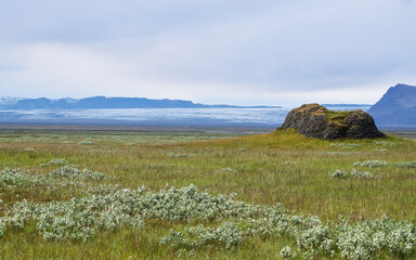 A grassy plain in southern Iceland leads to a solitary lava outcrop set against the distant...