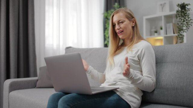 Attractive lady in domestic clothing having video conference on wireless laptop while sitting on couch. Happy blonde having online conversation at home.