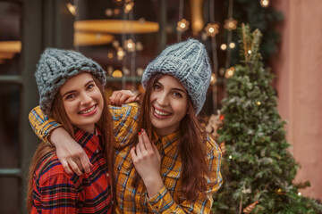 Christmas, New Year, winter holidays conception: two happy smiling women, friends, sisters, wearing knitted hat, tartan shirts, posing in street with festive decorations. Copy, empty space for text